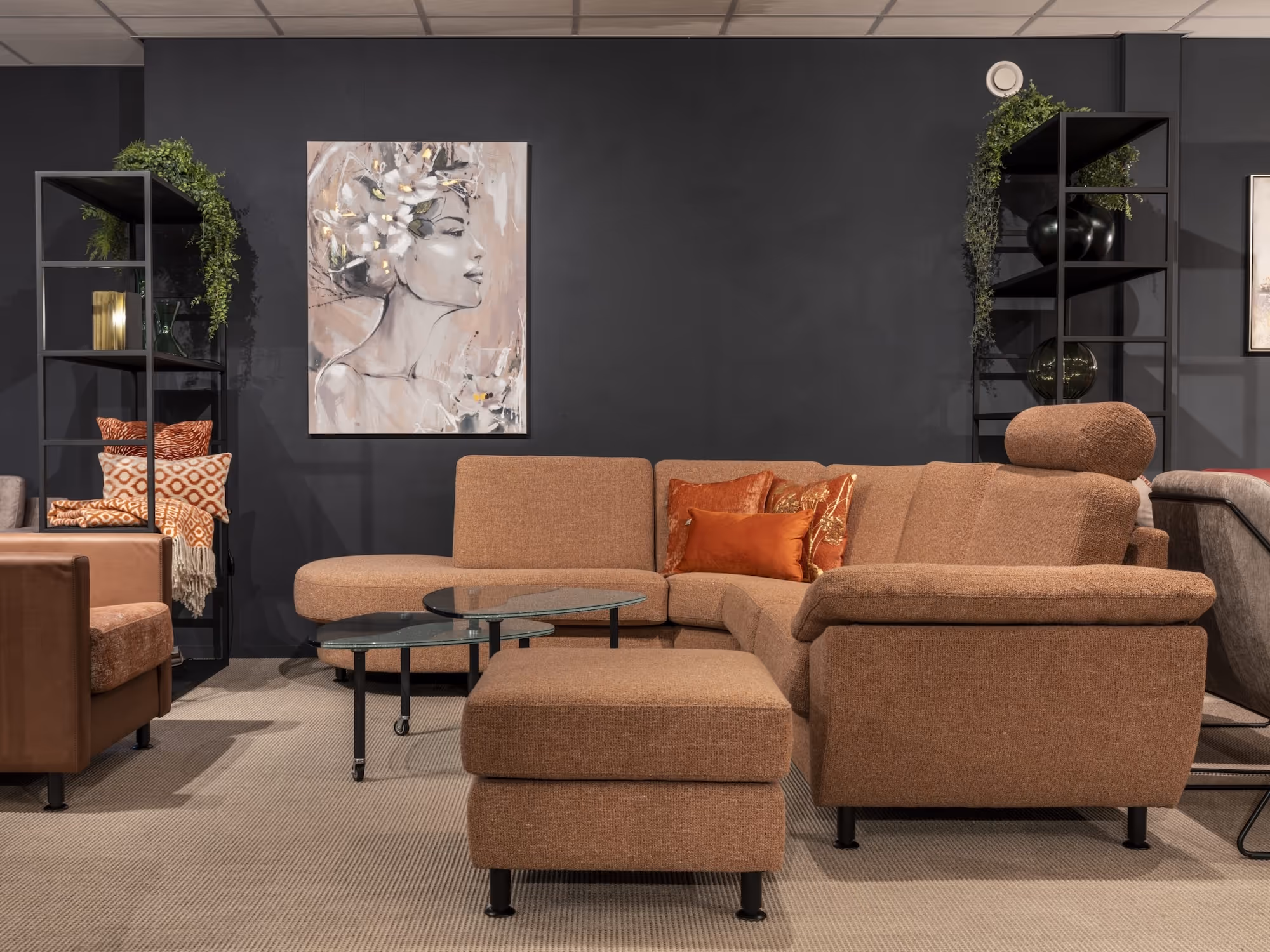 Modern living room with brown sectional sofa, orange throw pillows, glass coffee tables, and decorative black shelves with plants and artwork.