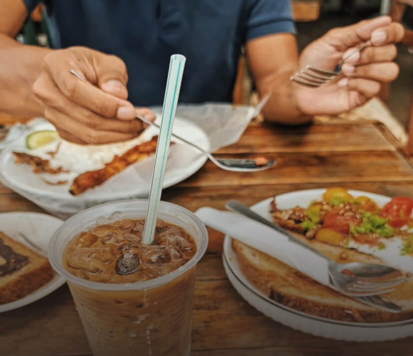 A person holding a fork eating a breakfast with toast, iced coffee, bacon and pastries
