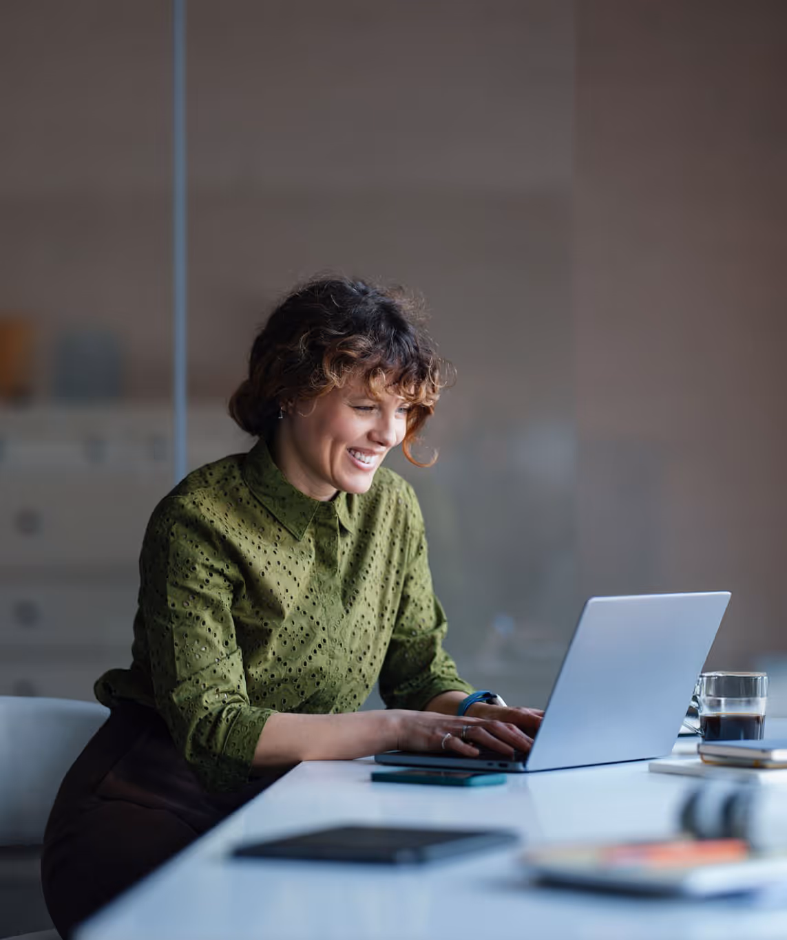 Smiling woman with curly hair in a green blouse typing on a laptop at a white table with a glass of coffee nearby.