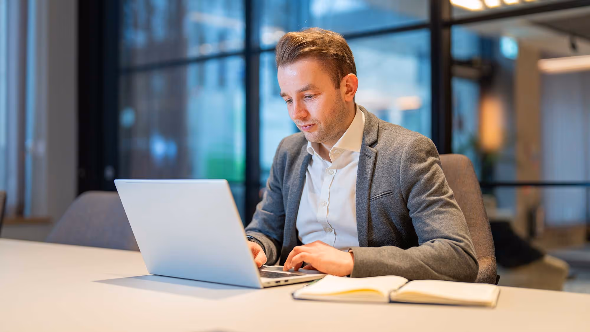 Ben Robinson in gray blazer working on a laptop at a desk with an open notebook in a modern office.