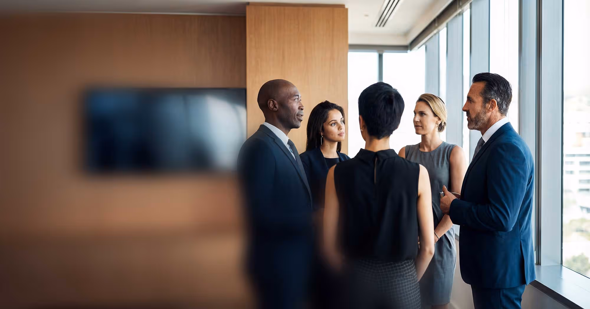 Five business professionals in formal attire having a discussion in a modern office with large windows.