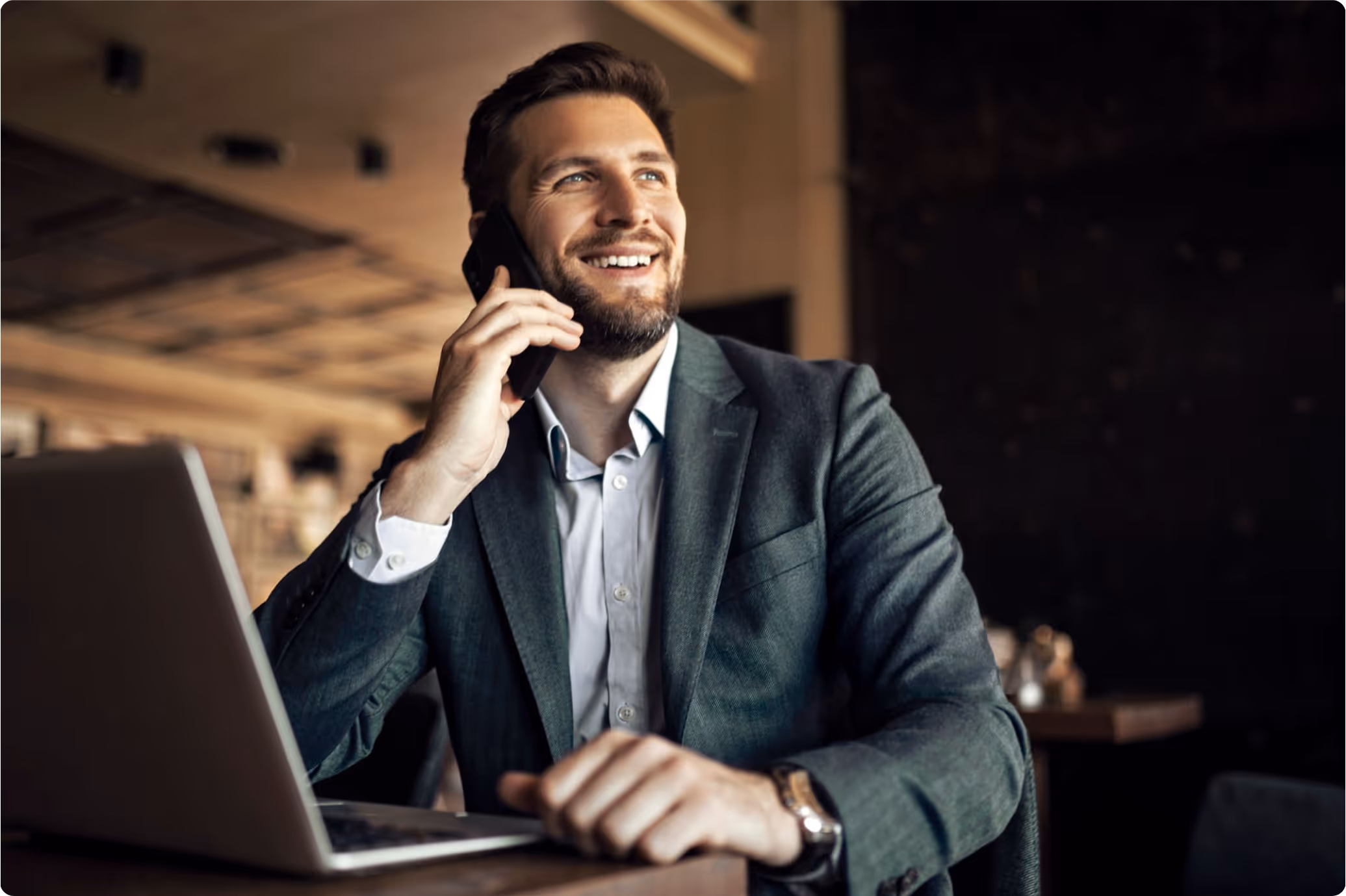 Smiling man in a suit talking on a smartphone while seated at a table with an open laptop.