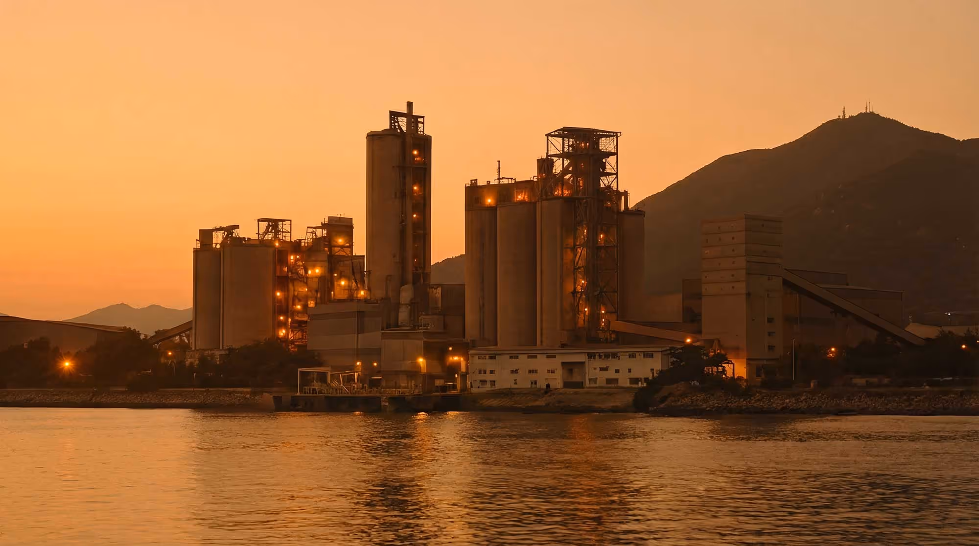 Industrial concrete plant with lit towers at sunset near calm water and silhouetted mountains.