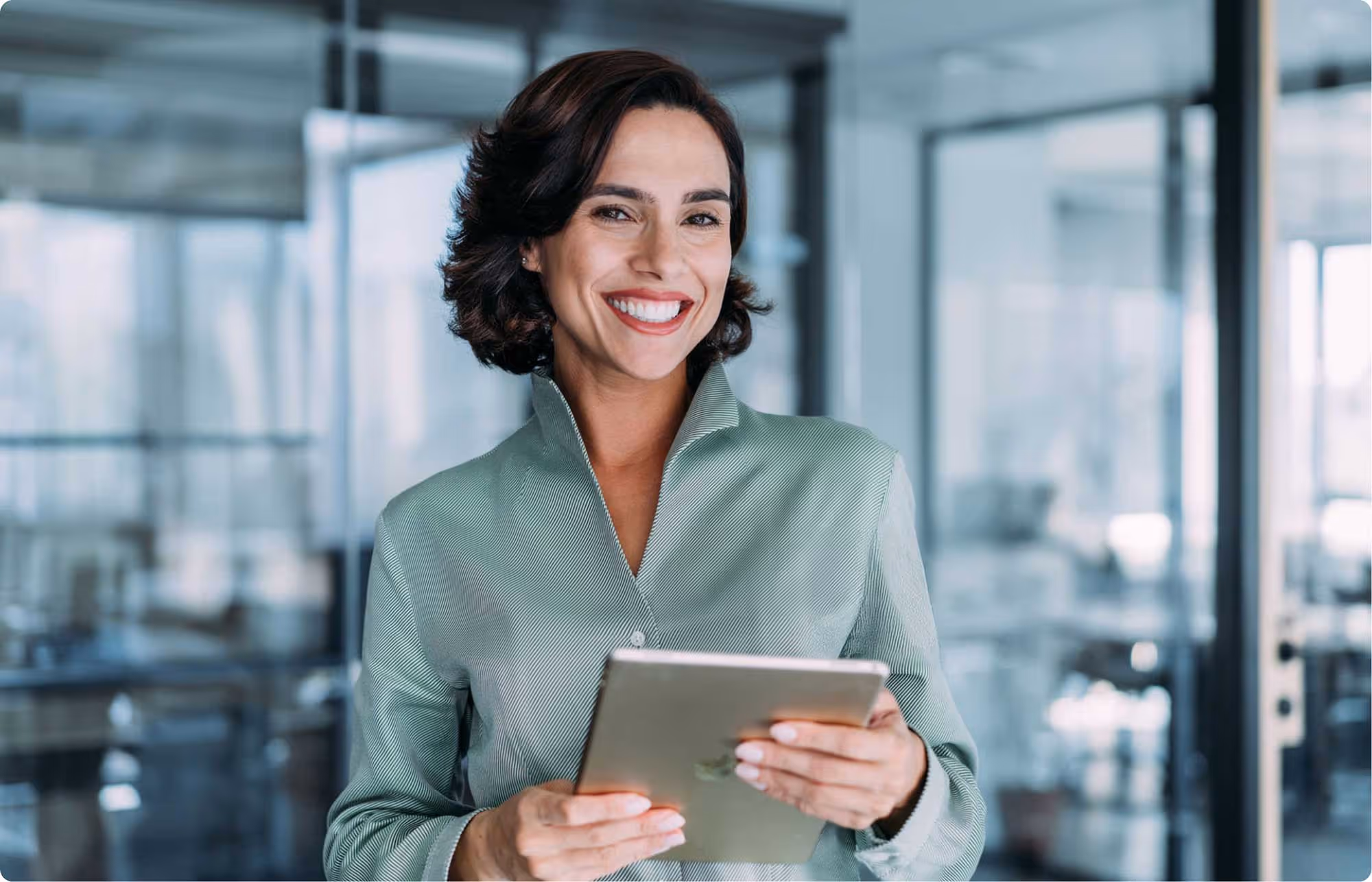 Smiling woman in a light green shirt holding a tablet in a modern office setting.