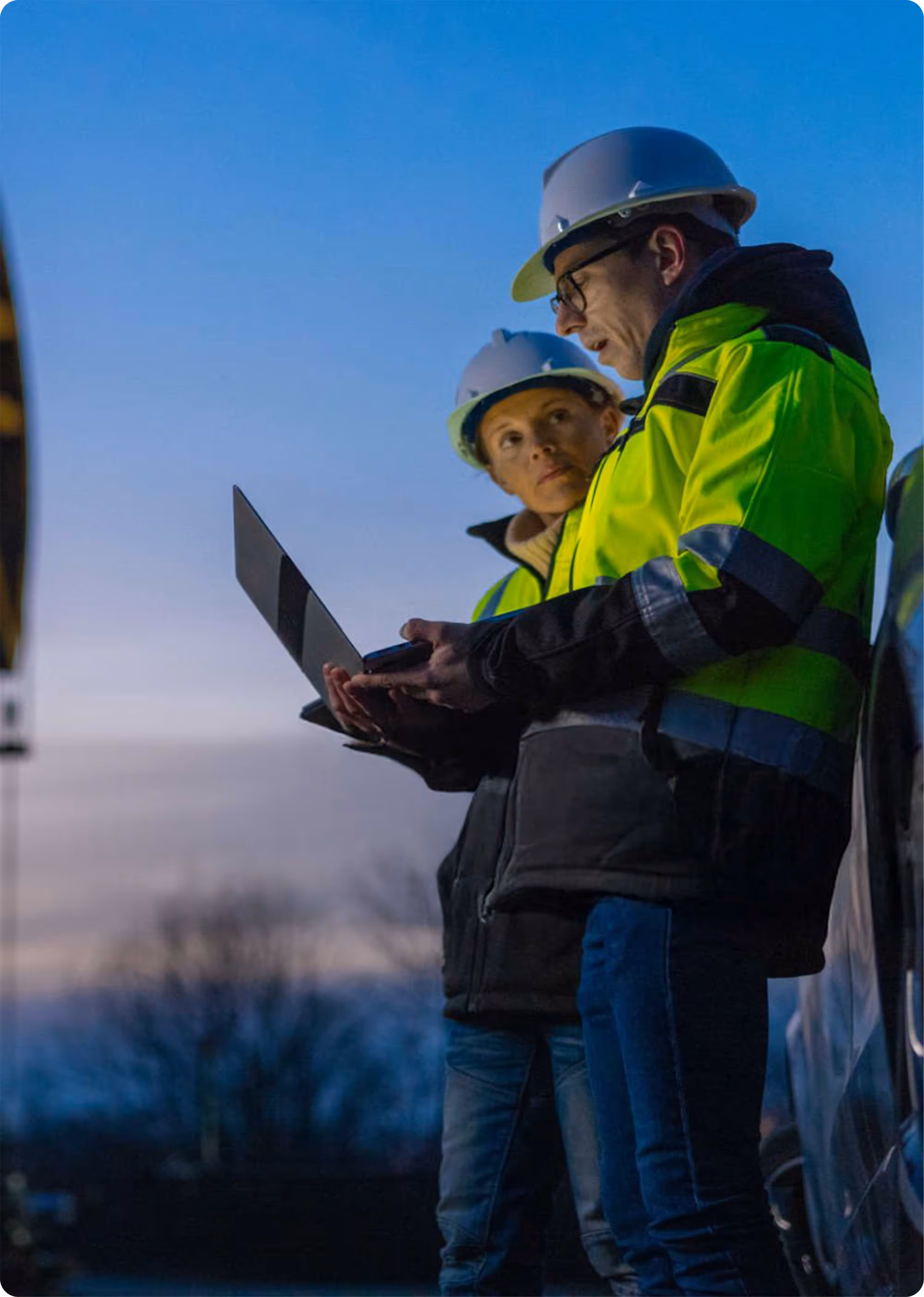 Two construction workers wearing yellow safety jackets and white helmets reviewing plans on a laptop outdoors at dusk.