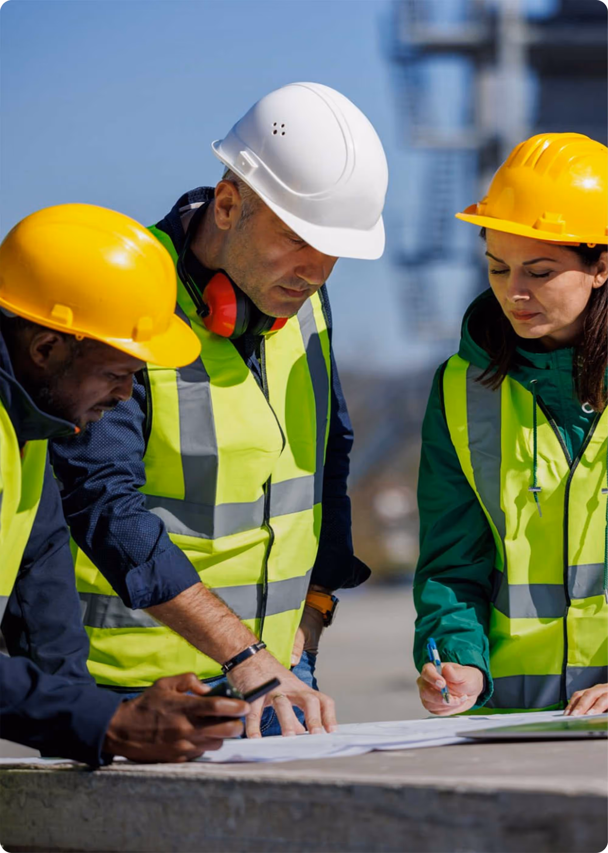 Three construction workers wearing yellow and white hard hats and reflective vests reviewing blueprints at a construction site.