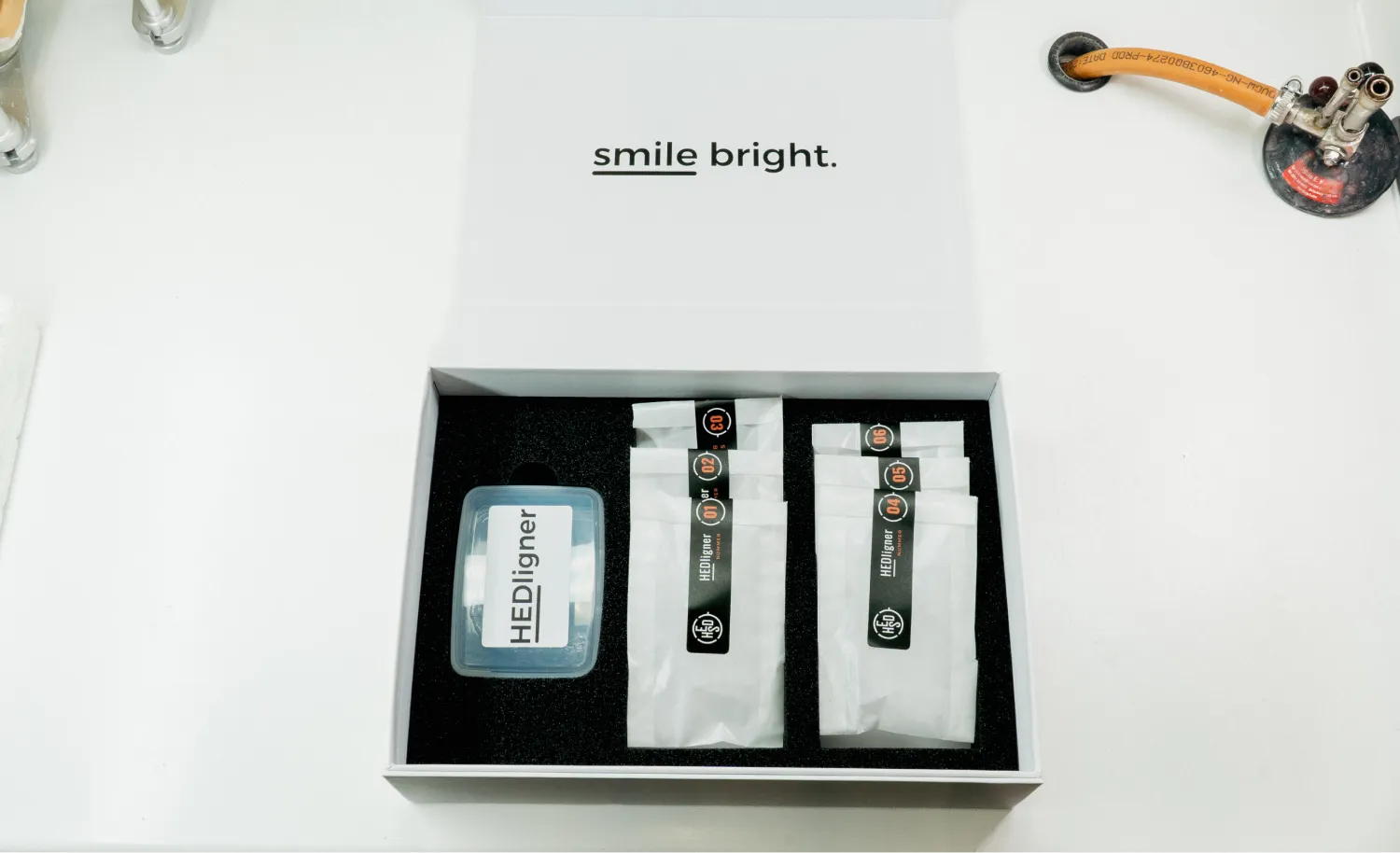 Weiße Box mit dem Text 'smile bright.' enthält einen transparenten Behälter mit der Aufschrift HEDiligner und sechs weiß verpackte Beutel mit Nummern von 01 bis 06.