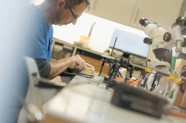 Techniker arbeitet konzentriert an einem kleinen mechanischen Gerät in einem Labor mit Mikroskopen und Werkzeugen.