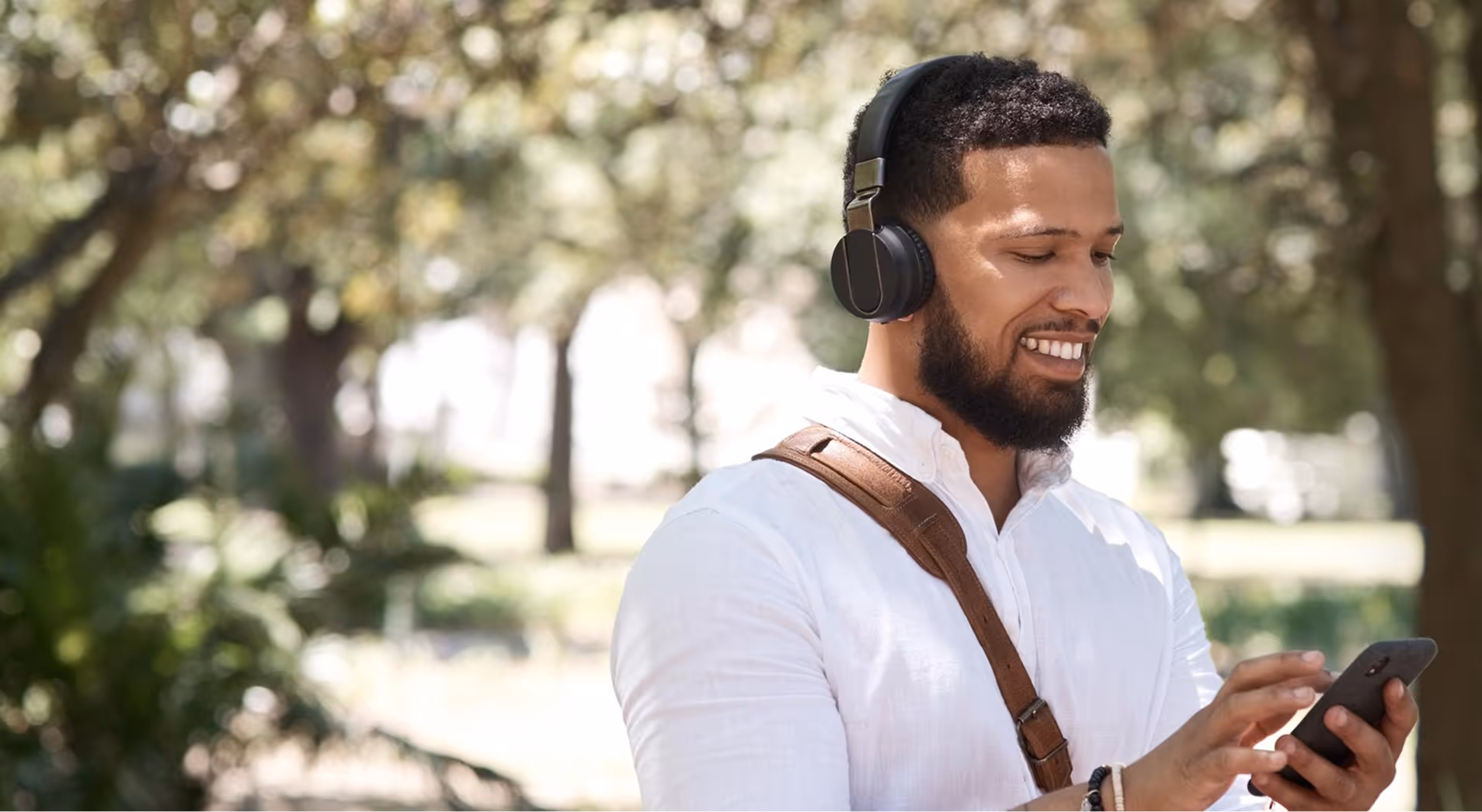 Smiling man with beard wearing black headphones and white shirt, using a smartphone outdoors with greenery in the background.