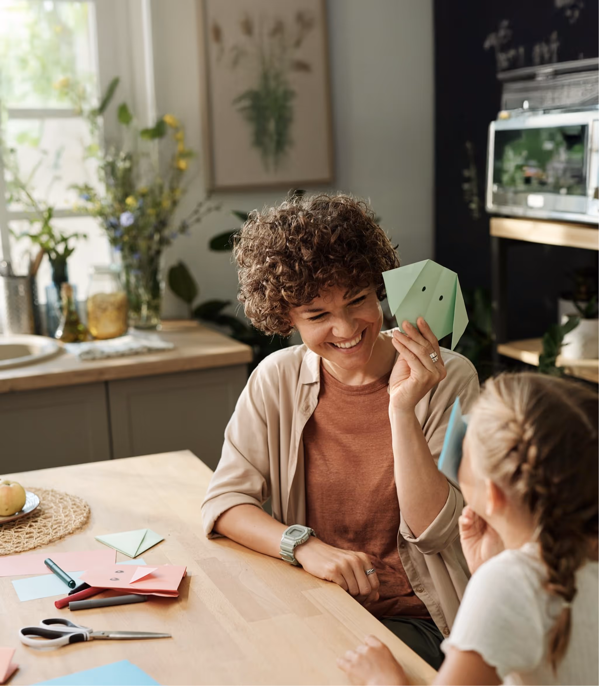 Curly-haired woman smiling and holding a green paper origami elephant while sitting at a table with a young girl.