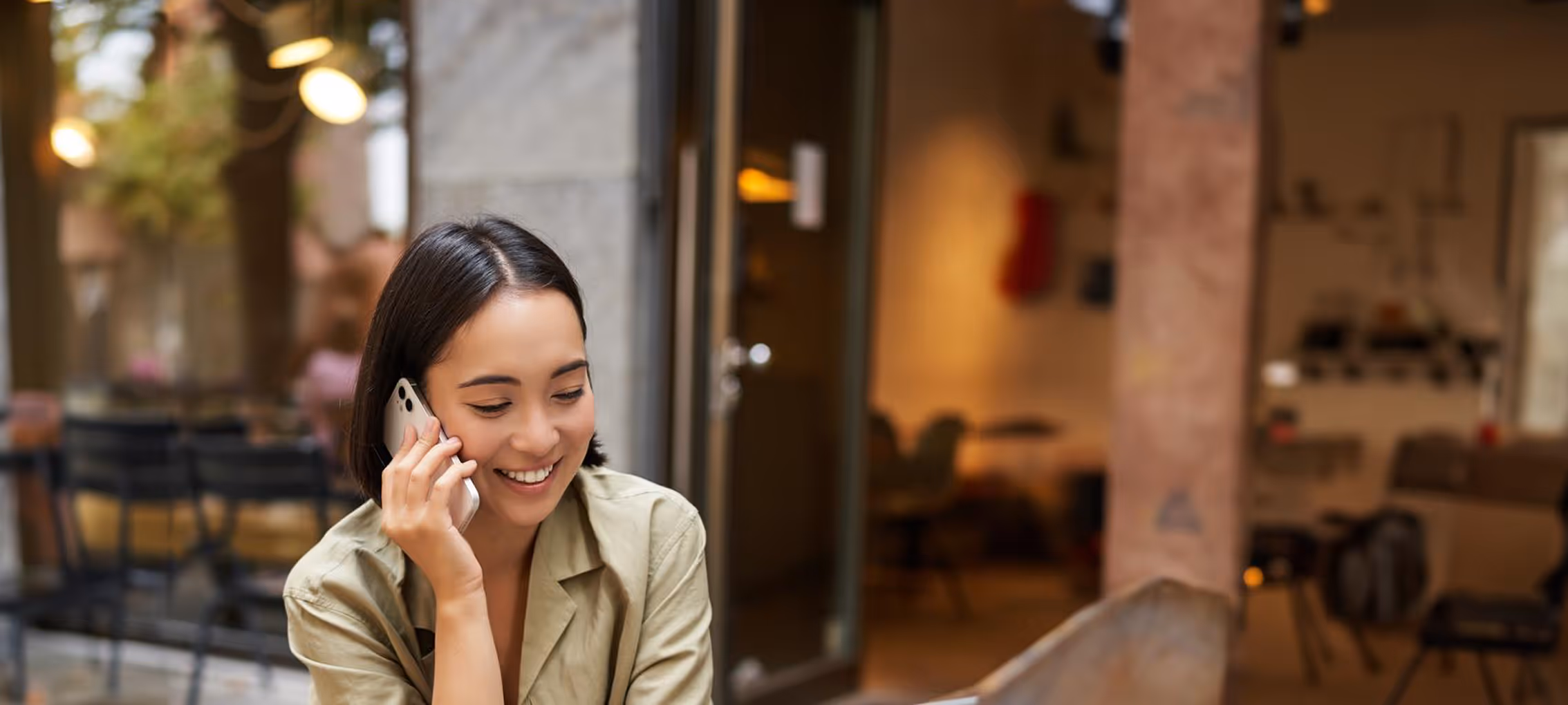 Smiling woman with short dark hair talking on a smartphone in an outdoor cafe setting.