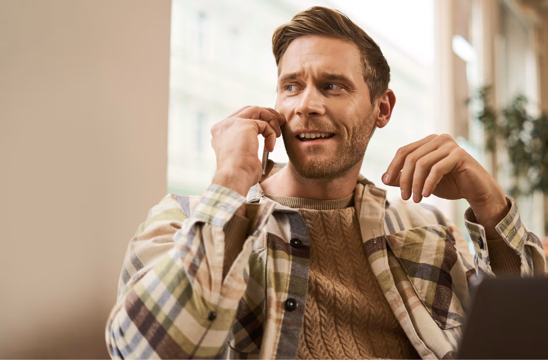 Man in a plaid shirt and brown sweater talking on a smartphone indoors with a window in the background.