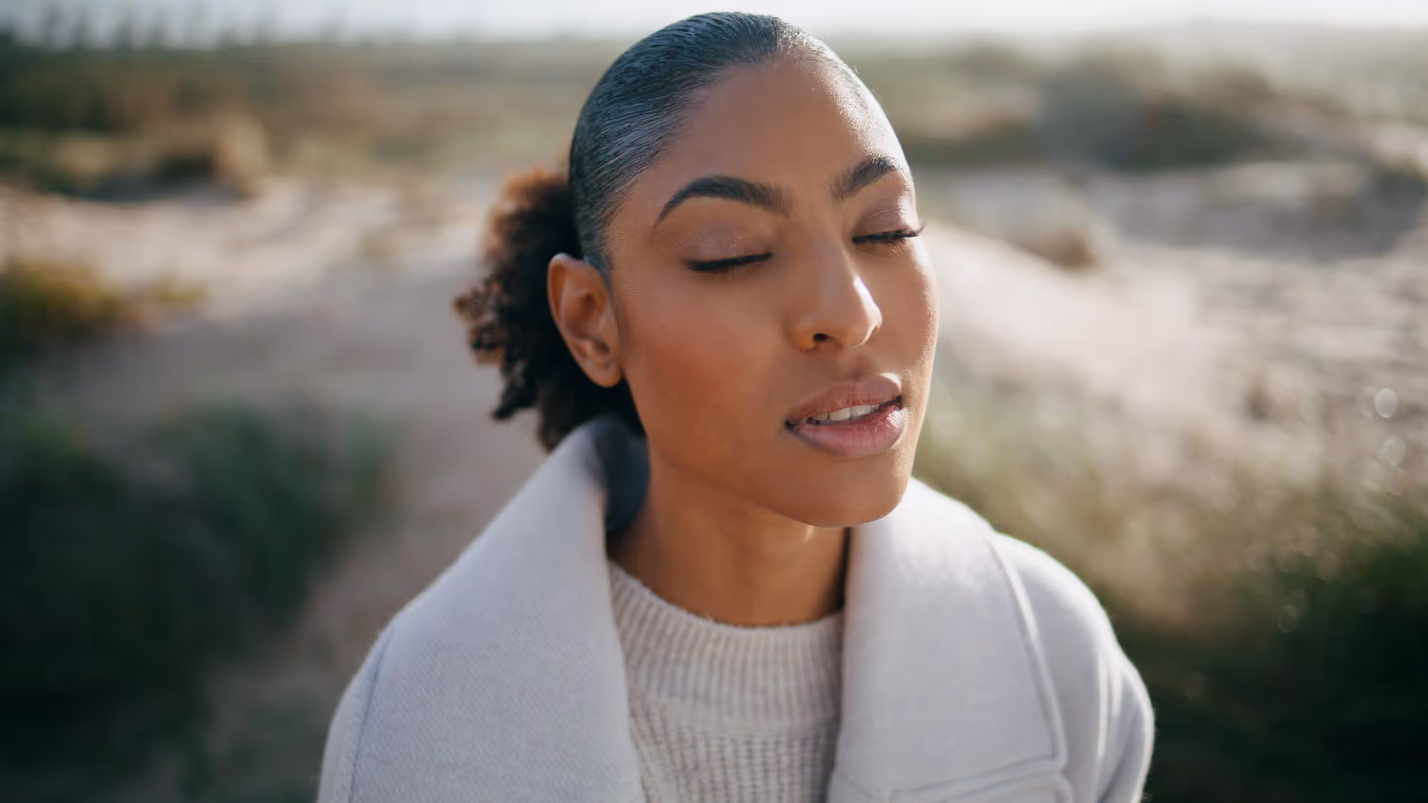 Close-up of a woman with eyes closed wearing a beige coat and sweater outdoors with a blurred natural background.
