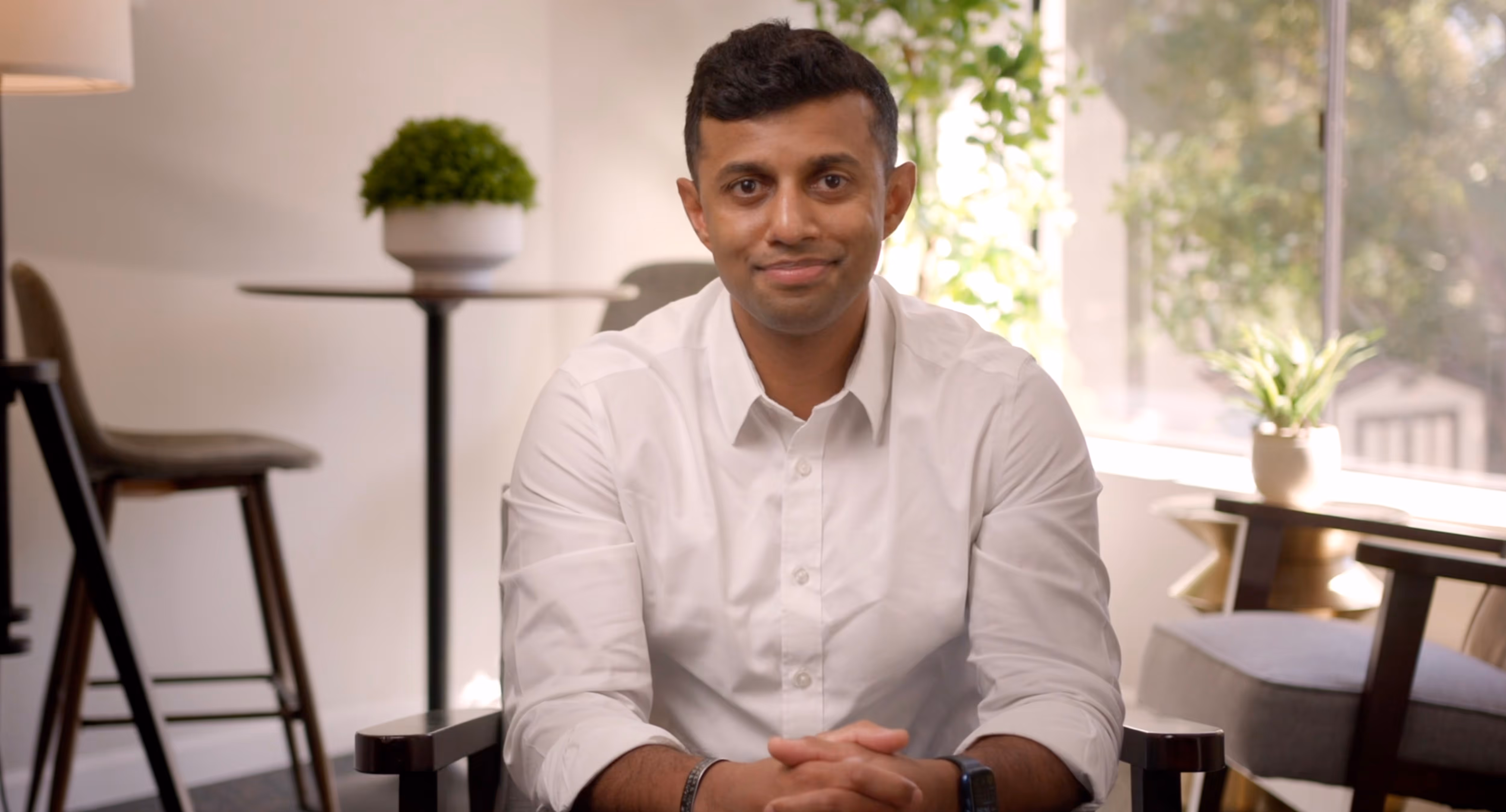 Smiling man in a white shirt sitting in a modern room with plants and chairs.