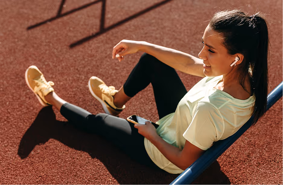 Young woman in sportswear with earbuds sitting and smiling on a track holding a smartphone.