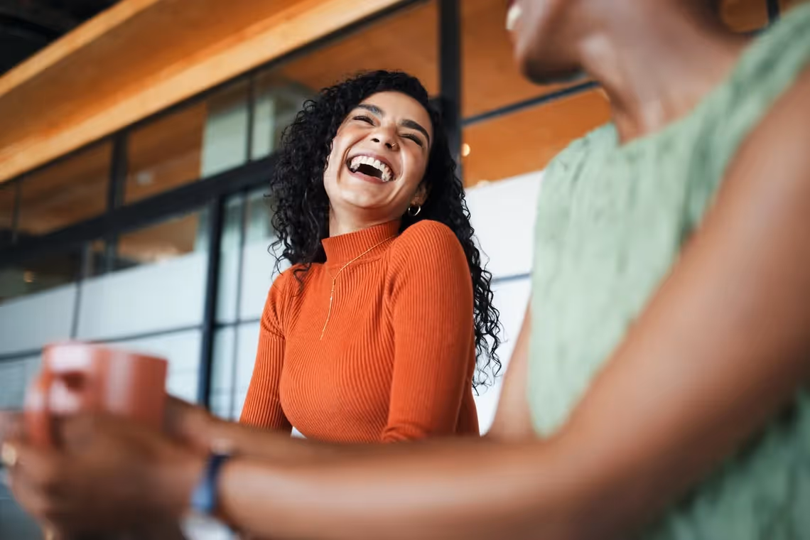 Woman in an orange sweater laughing joyfully while talking with another person holding a coffee cup.