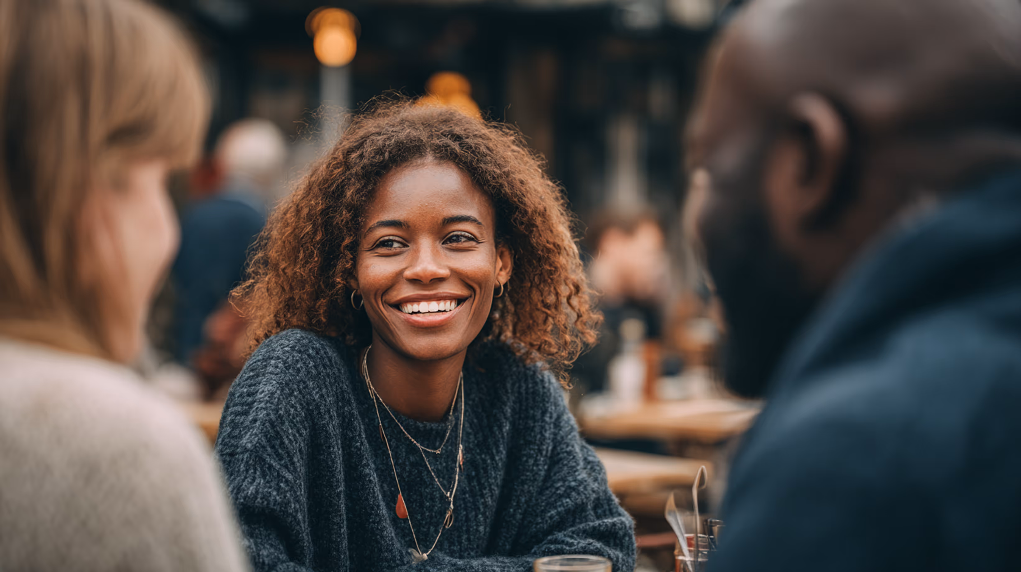 Smiling young woman with curly hair talking with two people at an outdoor café.