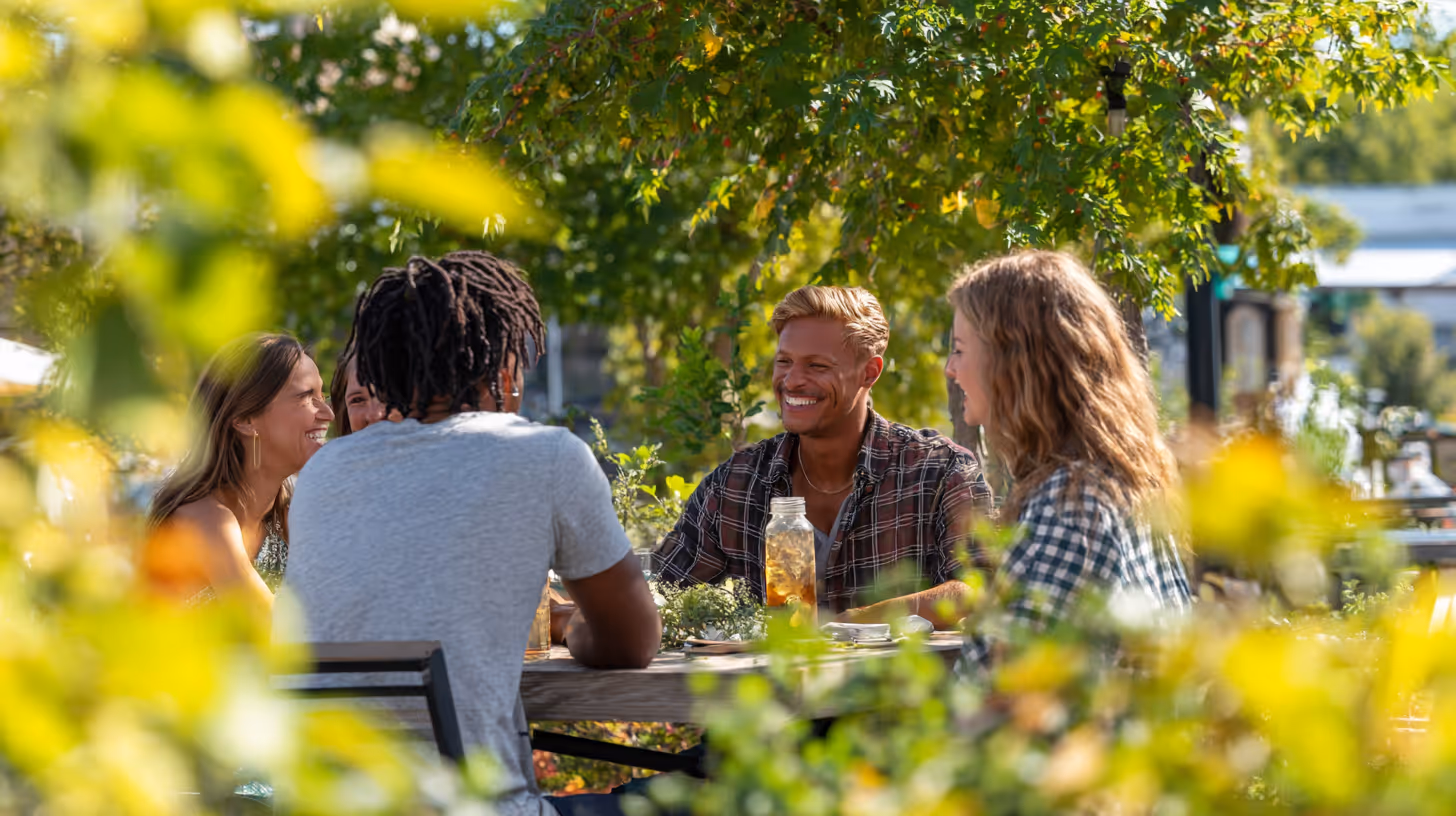 Group of four friends sitting at an outdoor table surrounded by green foliage, smiling and talking in the sunlight.