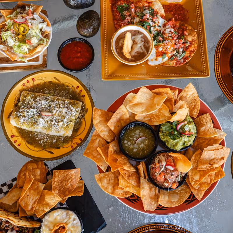 Table with Mexican dishes including a plate of tortilla chips with salsa, guacamole, and pico de gallo, a green sauce-covered enchilada, tostada salad, fried taquitos, and avocado halves.