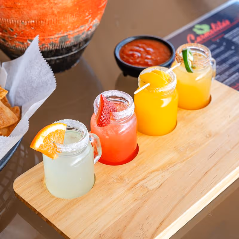 Four small mason jar cocktails garnished with orange, strawberry, peach, and jalapeño slices on a wooden serving board, with a side of chips and salsa.