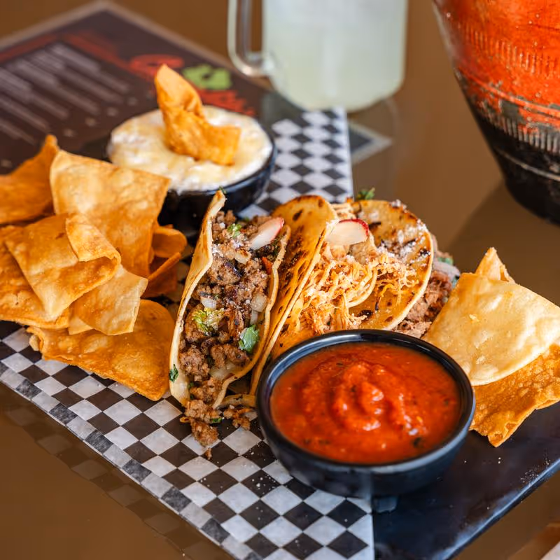 Plate with three tacos filled with ground meat, onions, cilantro, and radish slices, served with tortilla chips and bowls of red salsa and cheese dip.