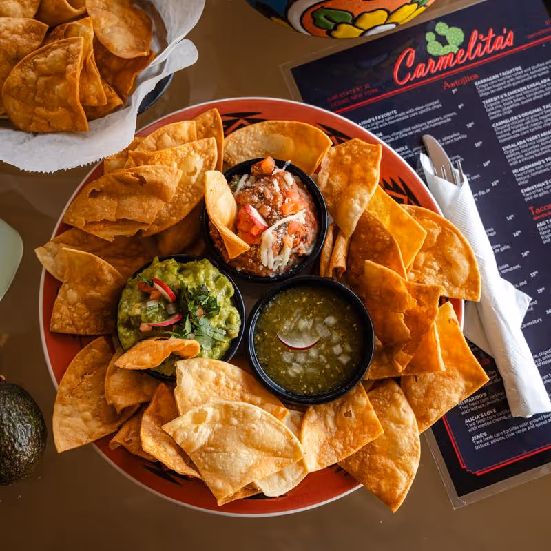 Plate of tortilla chips served with bowls of guacamole, salsa, and green sauce on a table next to a menu and wrapped utensils.