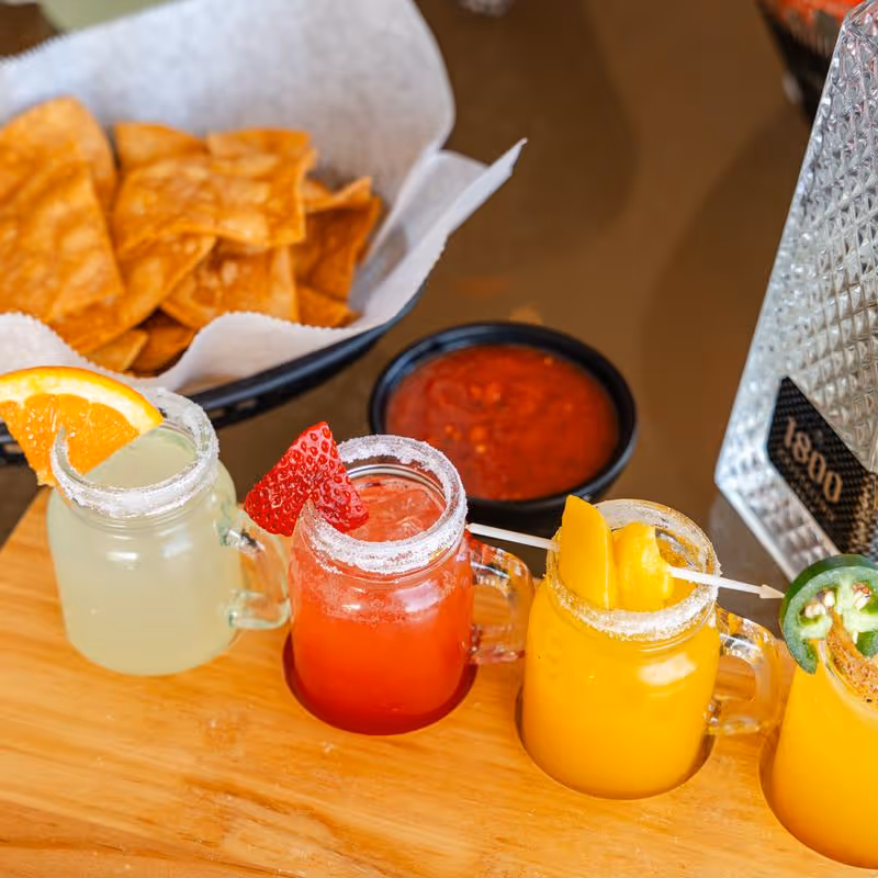 Four colorful cocktails garnished with orange, strawberry, mango, and jalapeño slices on a wooden board next to a basket of tortilla chips and a bowl of red salsa.