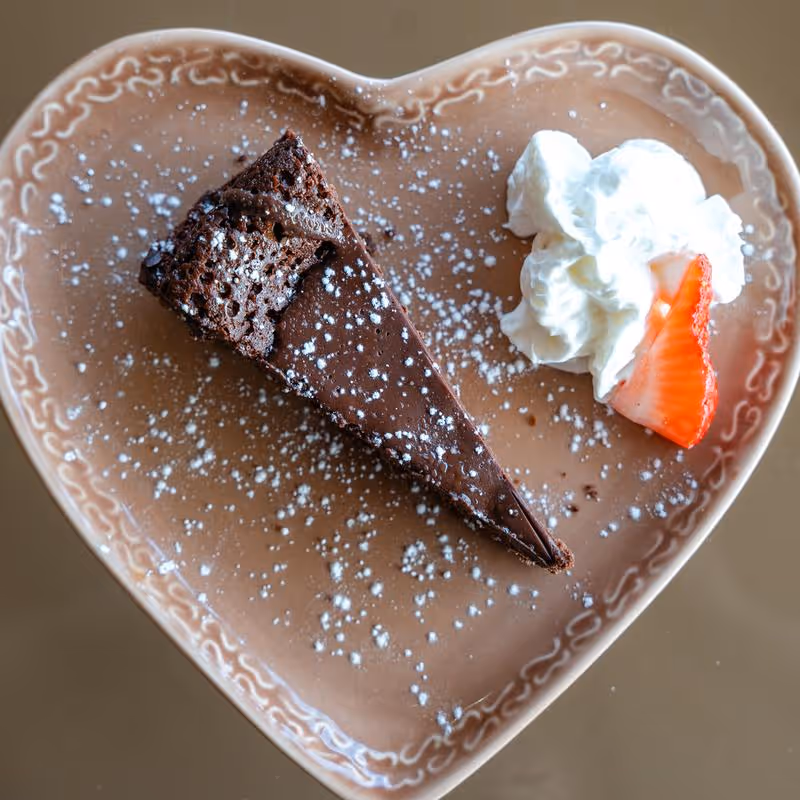 Slice of chocolate cake dusted with powdered sugar on a heart-shaped plate with whipped cream and a strawberry slice.