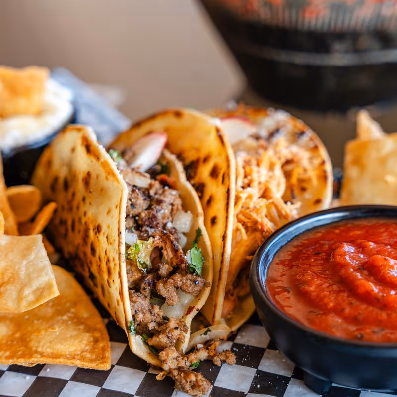 Close-up of two grilled tacos filled with seasoned meat, onions, and cilantro, served with a bowl of red salsa and tortilla chips on a checkered plate.