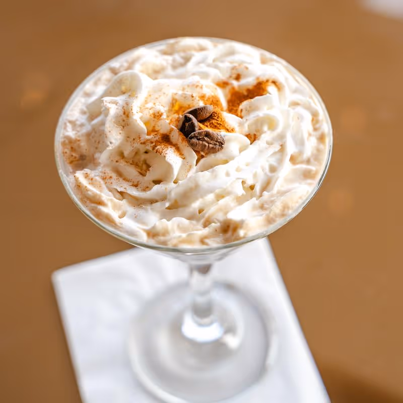 Close-up of a creamy coffee drink topped with whipped cream, cinnamon, and two coffee beans in a clear glass on a white napkin.