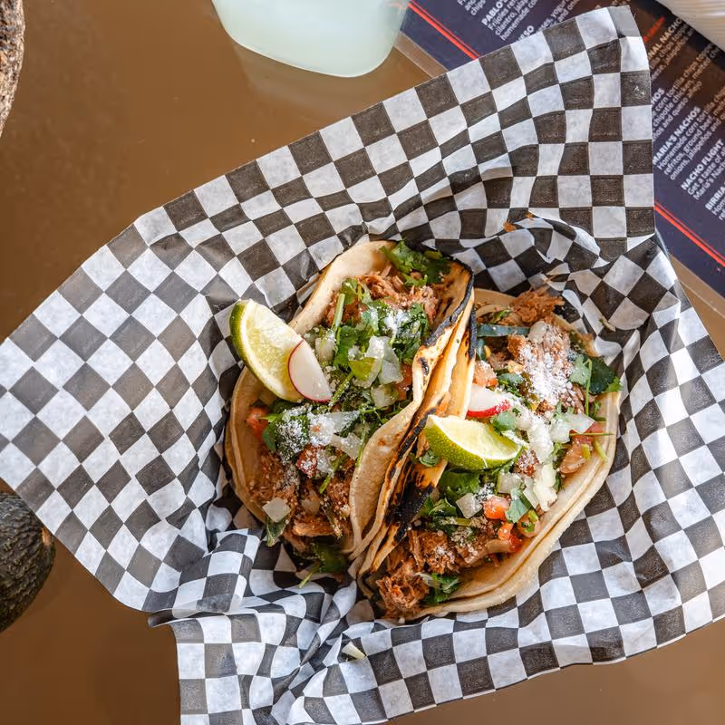 Two street tacos with shredded meat, chopped onions, cilantro, radish slices, cotija cheese, and lime wedges on checkered paper.