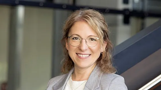 Frau mit Brille und hellgrauem Blazer lächelt vor unscharfem Bürohintergrund.