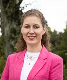 Frau mit lockigem Haar trägt pinkes Jackett, lächelt vor Baum und bewölktem Himmel.
