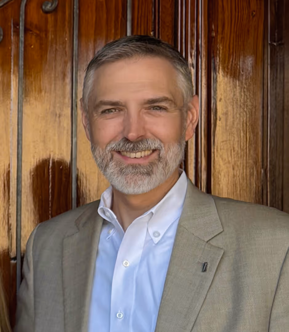 A photo of a bearded man standing in front of a wood door.