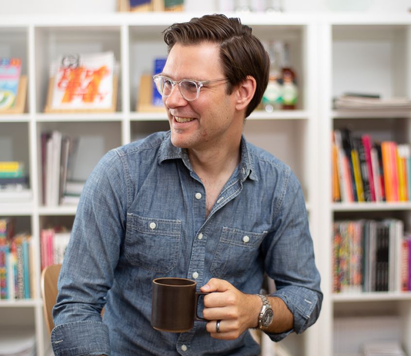 Man wearing glasses and a denim shirt, smiling and holding a brown mug while seated in front of a bookshelf.