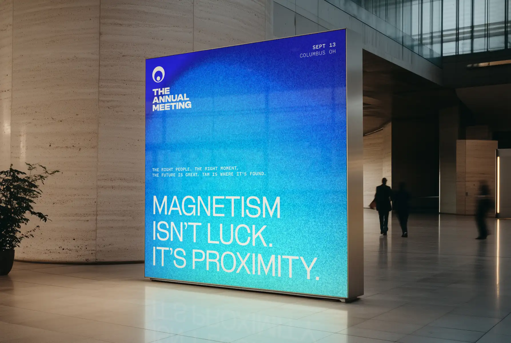 Large blue promotional display in a modern lobby reading 'The Annual Meeting - Magnetism isn't luck. It's proximity.' with date and location Sept 13 Columbus OH.