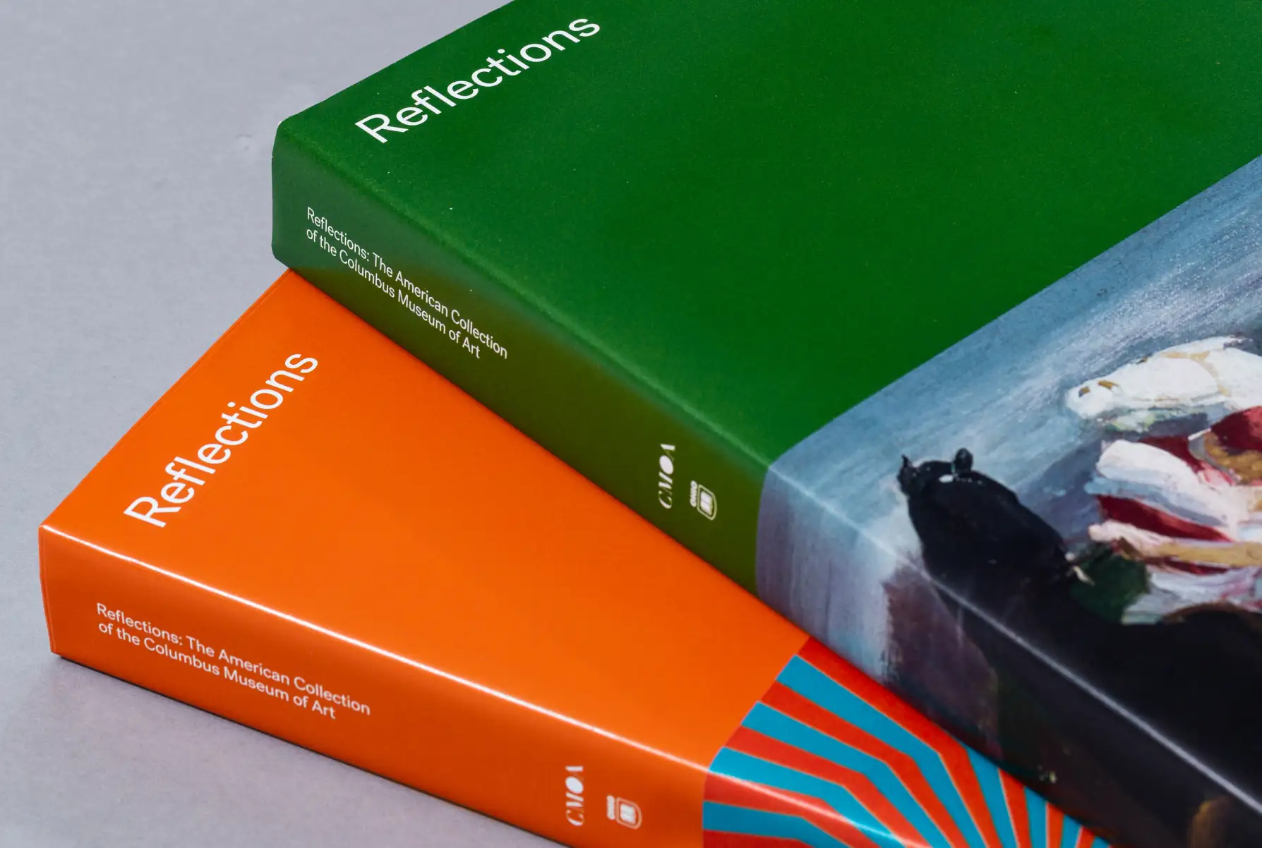 Two hardcover books titled 'Reflections: The American Collection of the Columbus Museum of Art' stacked, one with a green cover and the other orange.