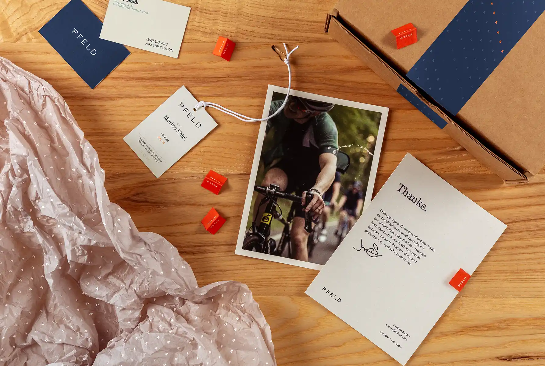 Flat lay of PFELD branded packaging including a thank you card with cyclist photo, product tag, business card, orange candies, and crumpled branded tissue paper on a wooden surface.