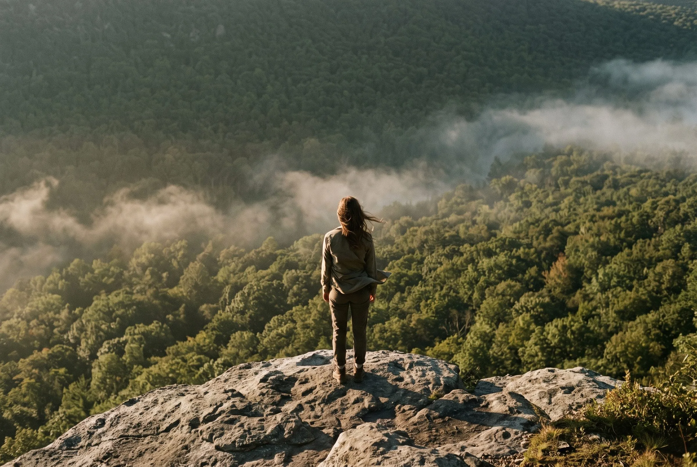 A woman standing on a rock ledge above a forested valley filled with morning cloud