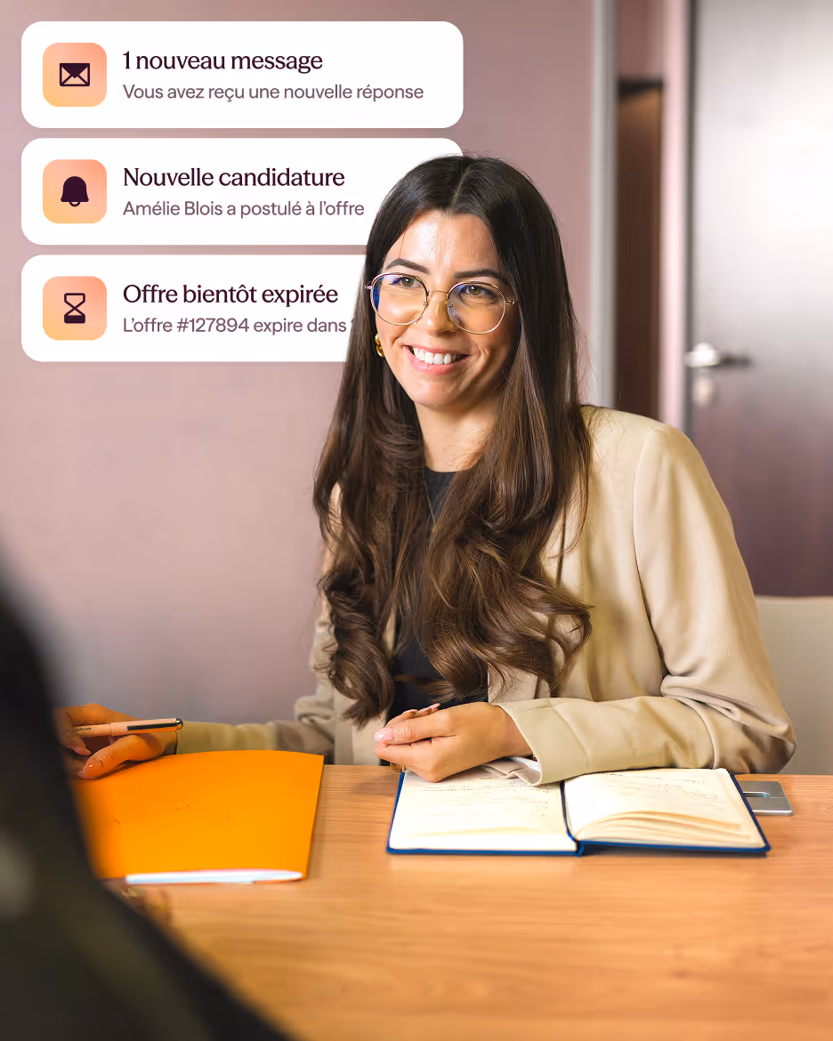 Femme souriante avec de longs cheveux bruns et des lunettes, assise à une table avec un carnet ouvert et un dossier orange, en entretien.