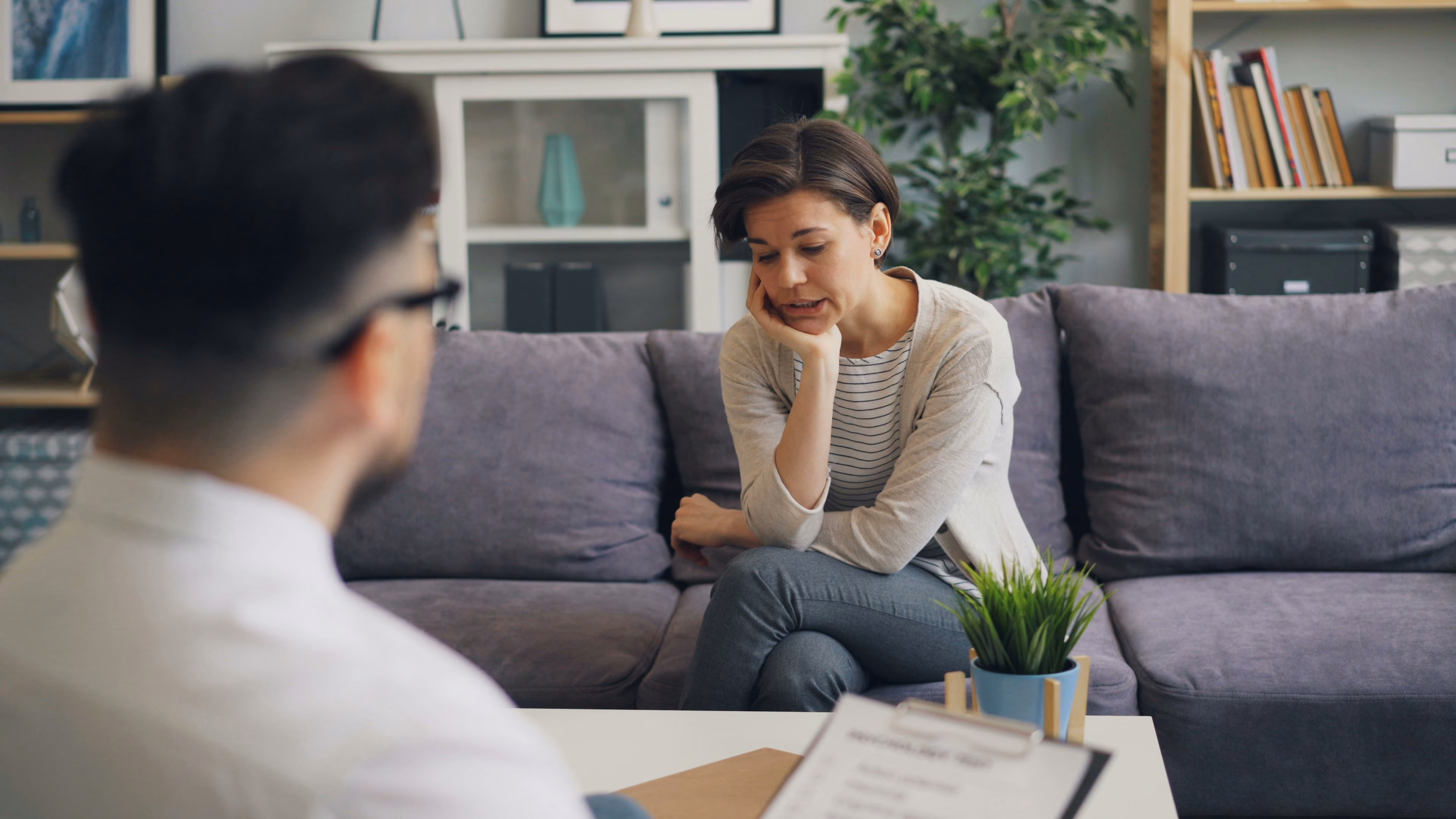 Woman sitting on couch, looking distressed, talking to a person holding a clipboard in a therapy session.