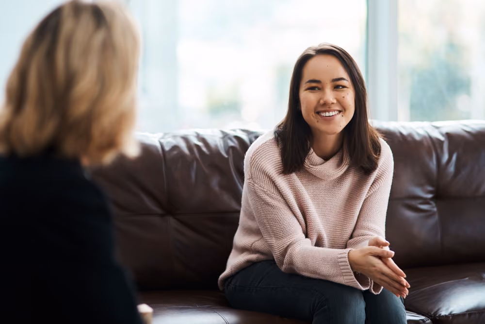 Smiling woman in a pink sweater sitting on a brown leather sofa during a conversation.