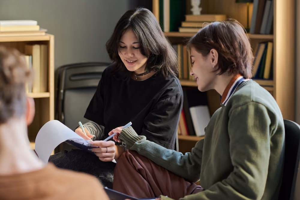 Two young women collaborate writing notes with bookshelves in the background.