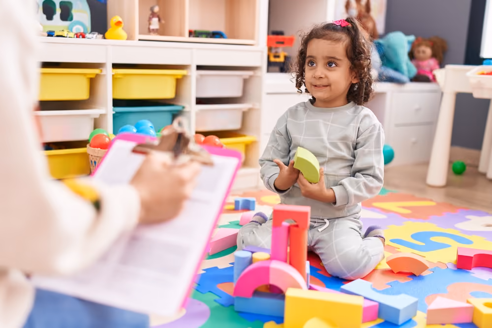 Child playing with colorful blocks on a puzzle mat while an adult takes notes on a clipboard.