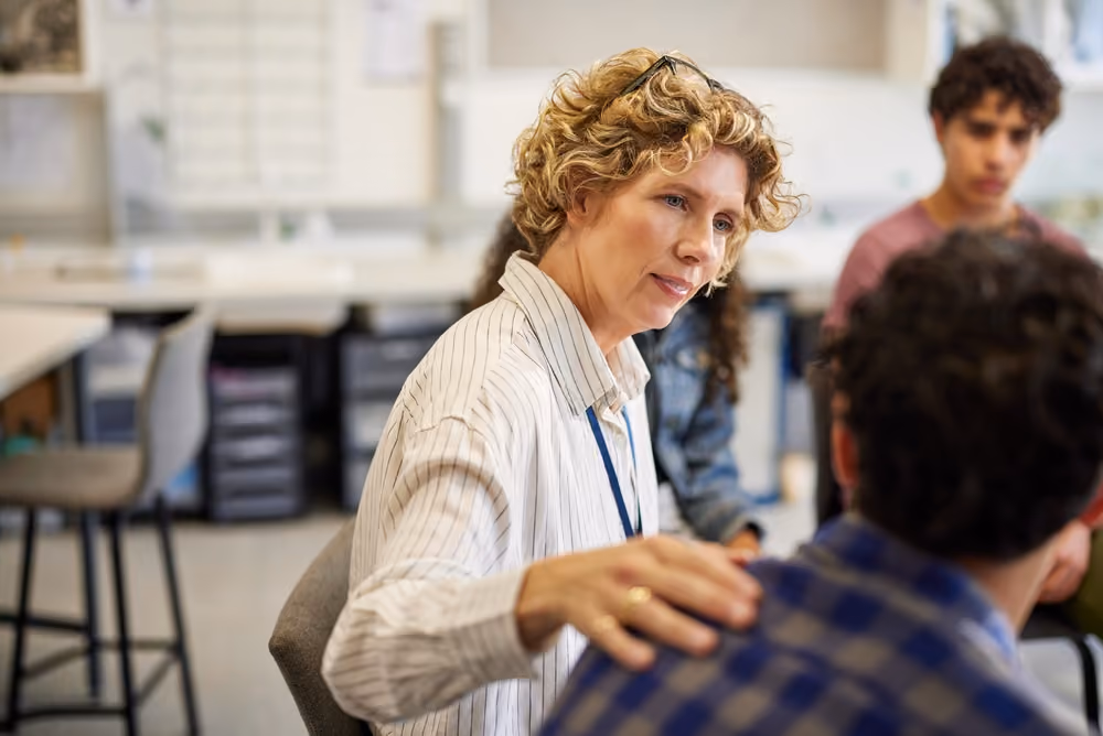 Woman with curly blonde hair comforting a seated person in a classroom setting.