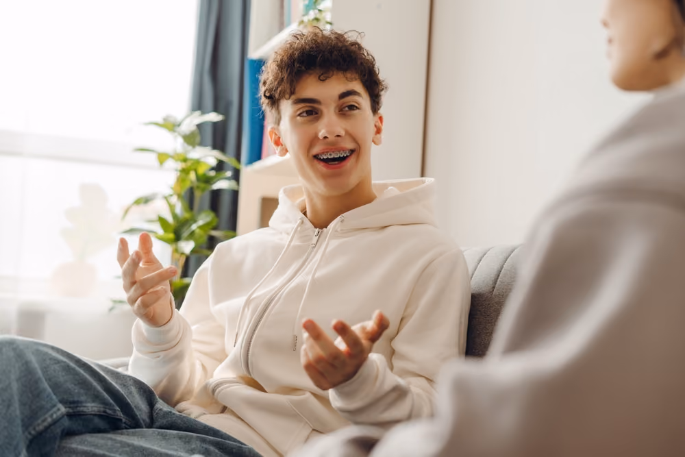 Teenage boy with braces wearing a white hoodie talking animatedly on a sofa indoors.