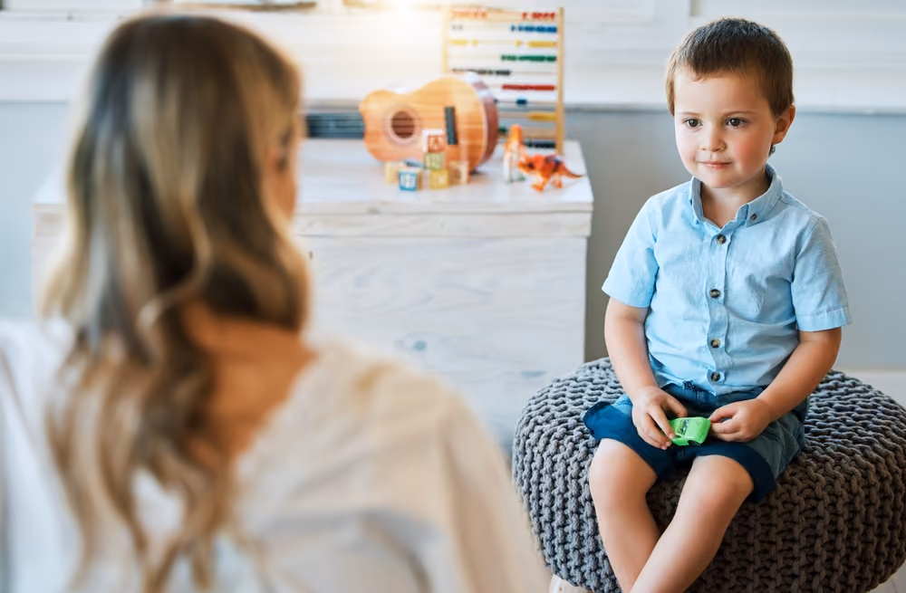 Young boy in blue shirt sitting on knitted ottoman holding a toy, facing a woman with long hair.