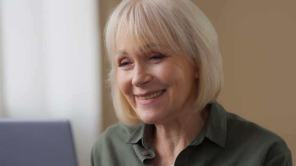 Smiling elderly woman with short blonde hair looking at a laptop screen.