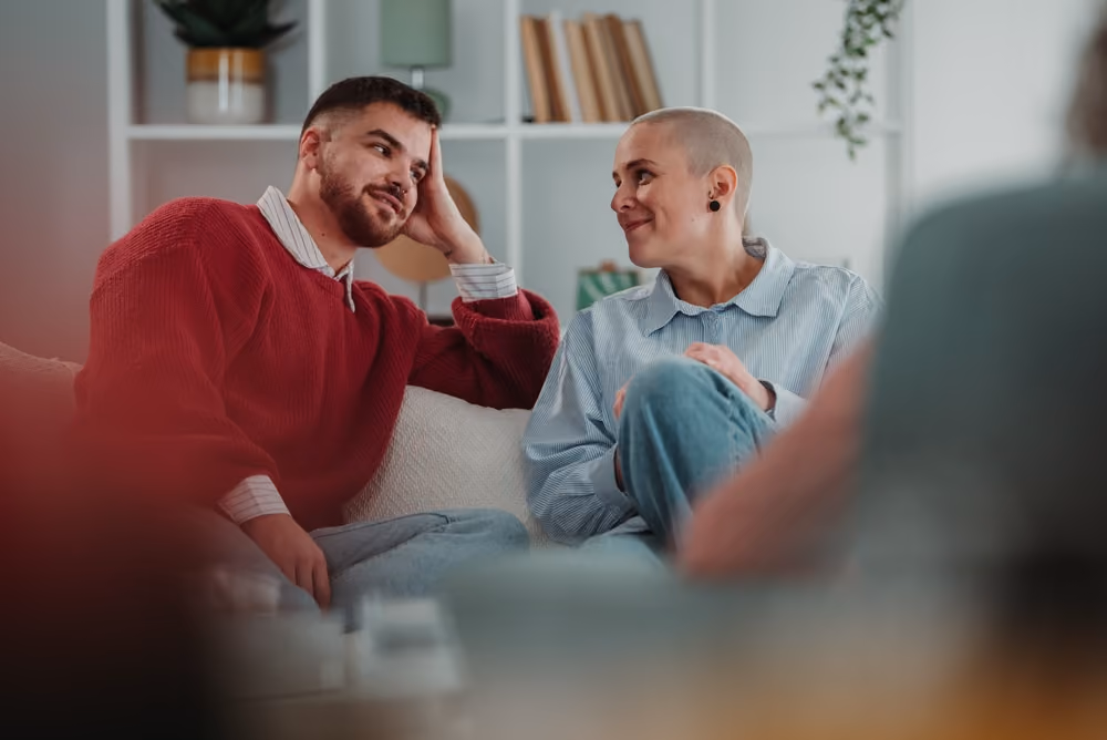 Two people sitting on a couch looking at each other and smiling in a cozy room.