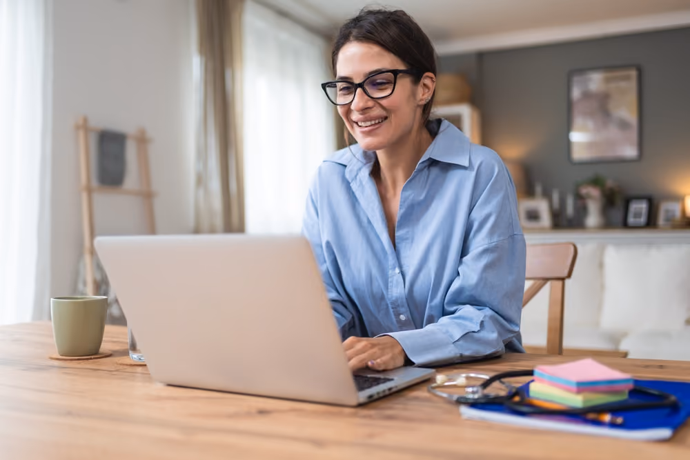 Smiling woman wearing glasses working on a laptop at a wooden table with a coffee mug nearby.